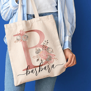 A person holds a tote bag with a large letter 'B', floral decorations, and the name \“Barbara\“ written in cursive. The person wears a striped blue shirt and jeans.