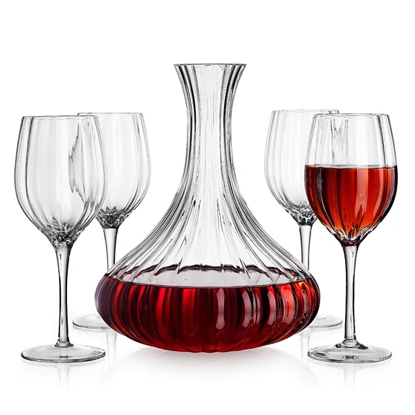 A clear glass wine decanter with a ribbed design filled with red wine is surrounded by four clear wine glasses, one of which contains wine.