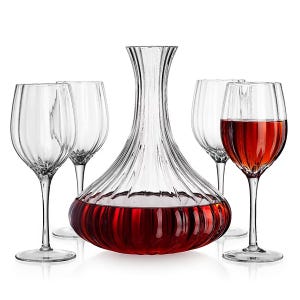 A clear glass wine decanter with a ribbed design filled with red wine is surrounded by four clear wine glasses, one of which contains wine.