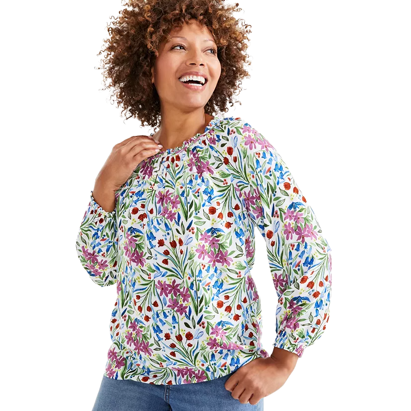 A woman is wearing a long-sleeve blouse with a colorful floral pattern in shades of purple, blue, red, and green. She pairs it with blue jeans and looks happy and relaxed.