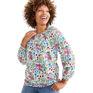 A woman is wearing a long-sleeve blouse with a colorful floral pattern in shades of purple, blue, red, and green. She pairs it with blue jeans and looks happy and relaxed.