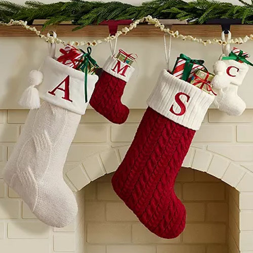 Four Christmas stockings with initials \“A,\“ \“M,\“ \“S,\“ and \“C,\“ hang by a fireplace, each filled with wrapped gifts. The stockings are in white, red, and cream cable knit designs.
