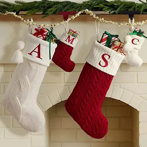 Four Christmas stockings with initials \“A,\“ \“M,\“ \“S,\“ and \“C,\“ hang by a fireplace, each filled with wrapped gifts. The stockings are in white, red, and cream cable knit designs.