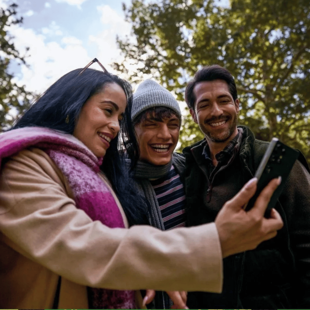 Three people are taking a selfie outdoors using a smartphone. They are dressed warmly and appear happy, suggesting a joyful moment shared among friends.
