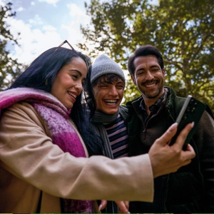 Three people are taking a selfie outdoors using a smartphone. They are dressed warmly and appear happy, suggesting a joyful moment shared among friends.
