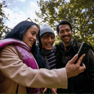 Three people are taking a selfie outdoors using a smartphone. They are dressed warmly and appear happy, suggesting a joyful moment shared among friends.