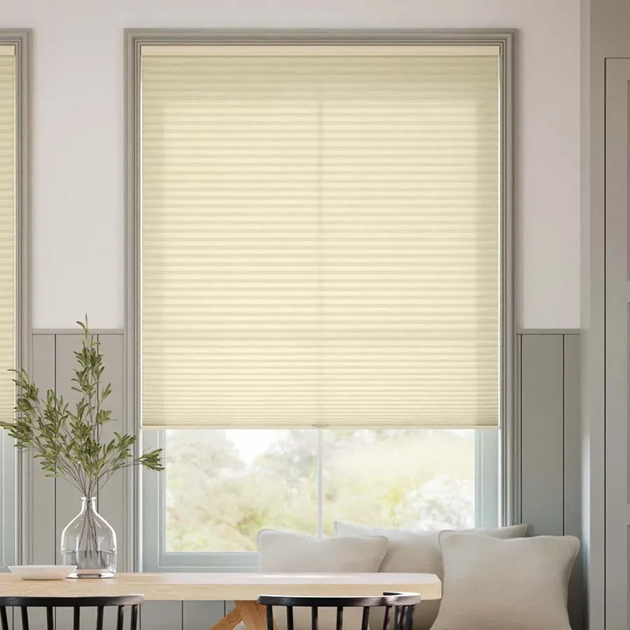 A window with cream-colored cellular blinds is partially drawn, complemented by a neutral-toned interior featuring a wooden table, chairs, a vase with greenery, and light-colored pillows.
