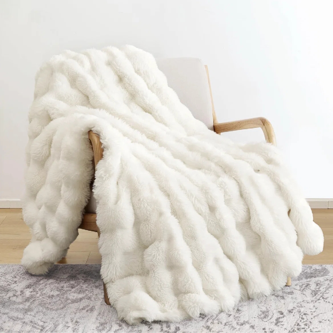 A white faux fur blanket with a textured, wavy pattern is draped over a wooden armchair on a light-patterned rug.