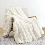 A white faux fur blanket with a textured, wavy pattern is draped over a wooden armchair on a light-patterned rug.