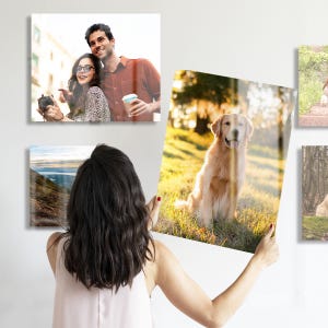 A woman is holding a large acrylic print of a dog, while other acrylic prints featuring a couple and a landscape are mounted on the wall. The prints have a glossy finish and showcase vibrant colors.