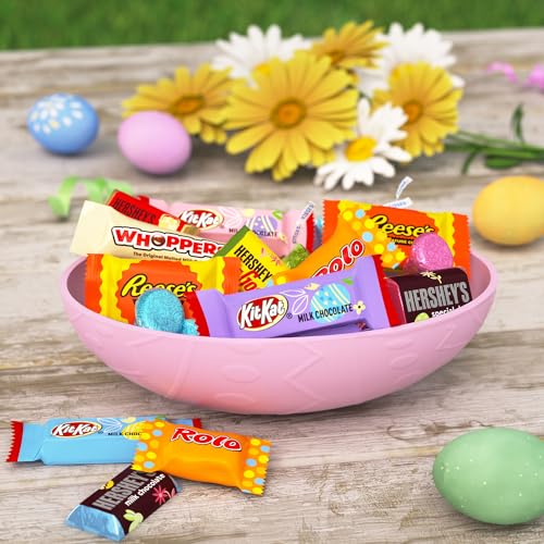 A pink Easter-themed bowl filled with assorted candies including Reese's, KitKat, Hershey's, Whoppers, and Rolo, set on a wooden surface amidst decorative Easter eggs and flowers.