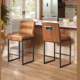 VASAGLE by SONGMICS HOME displays two brown leather bar stools with black metal frames in a kitchen setting, featuring a pocket on the backrest.