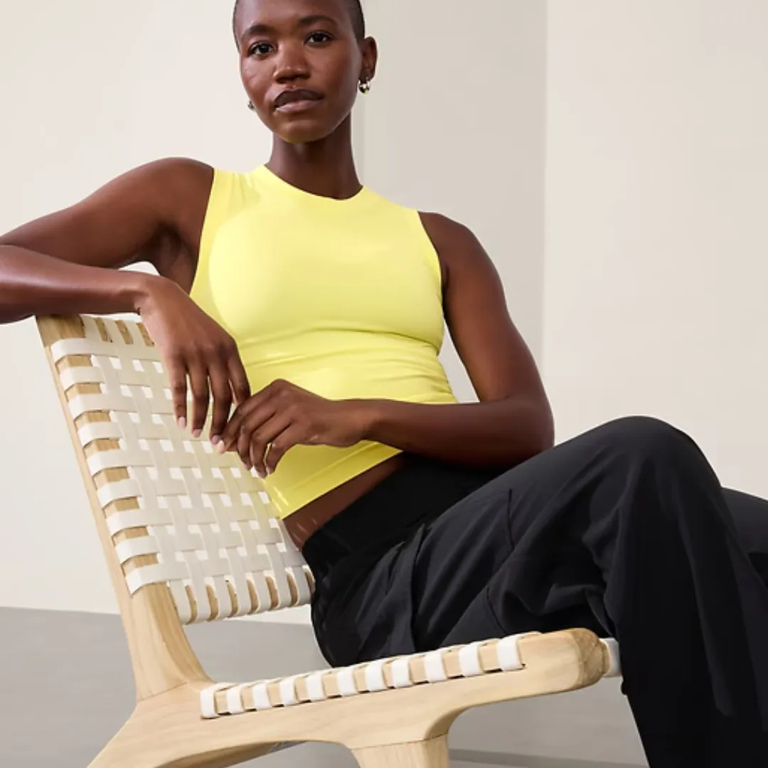 A woman is wearing a sleeveless yellow top and black trousers, seated on a wooden chair with white straps.