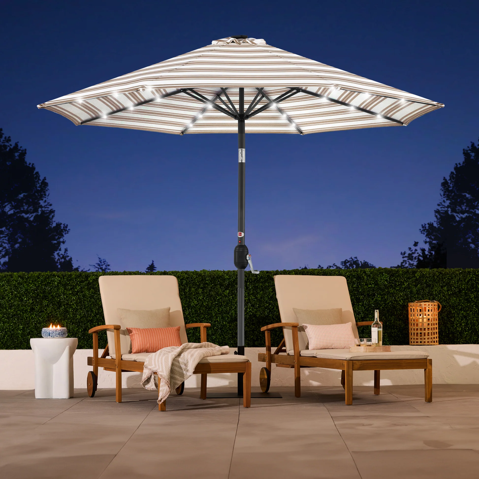 Two wooden lounge chairs with cushions and striped pillows sit beneath a large striped patio umbrella with LED lights, and nearby are a small round table with a candle and a bottle of wine.