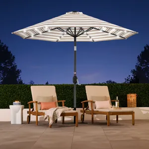 Two wooden lounge chairs with cushions and striped pillows sit beneath a large striped patio umbrella with LED lights, and nearby are a small round table with a candle and a bottle of wine.