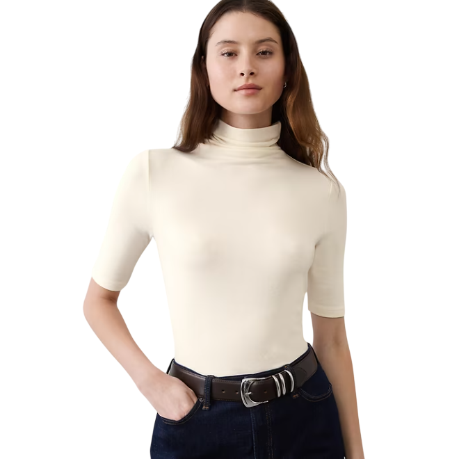 A woman wears a short-sleeve beige turtleneck top paired with dark blue jeans and a dark belt.