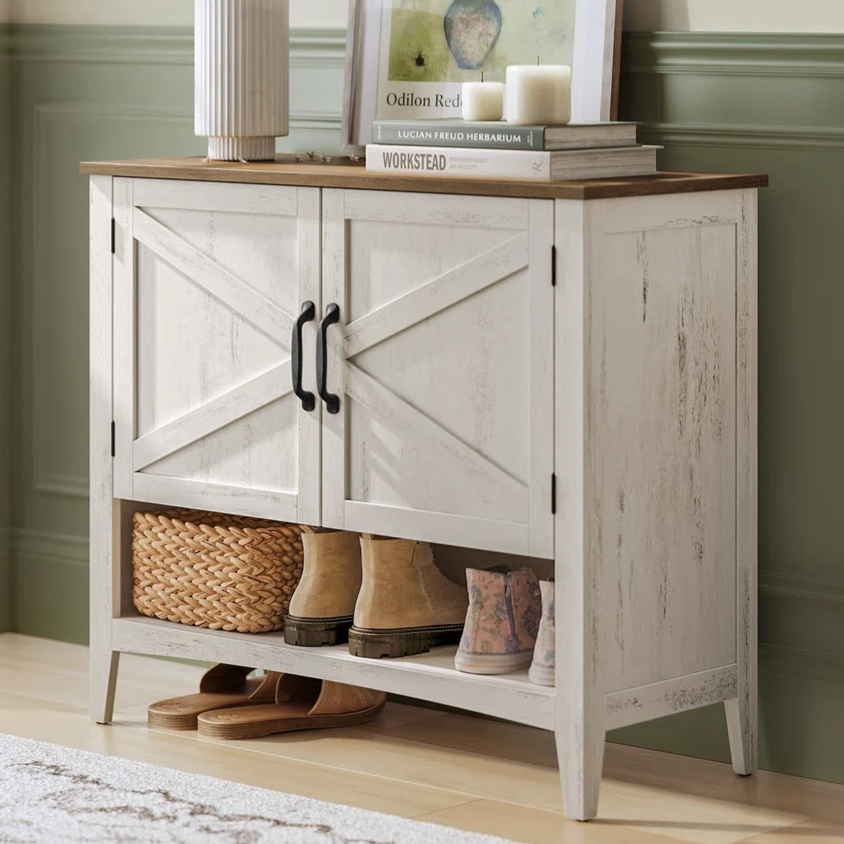 A rustic white cabinet with black handles, featuring two doors and an open lower shelf with shoes and a woven basket. The top holds decorative items including a vase with branches, framed art, and books.