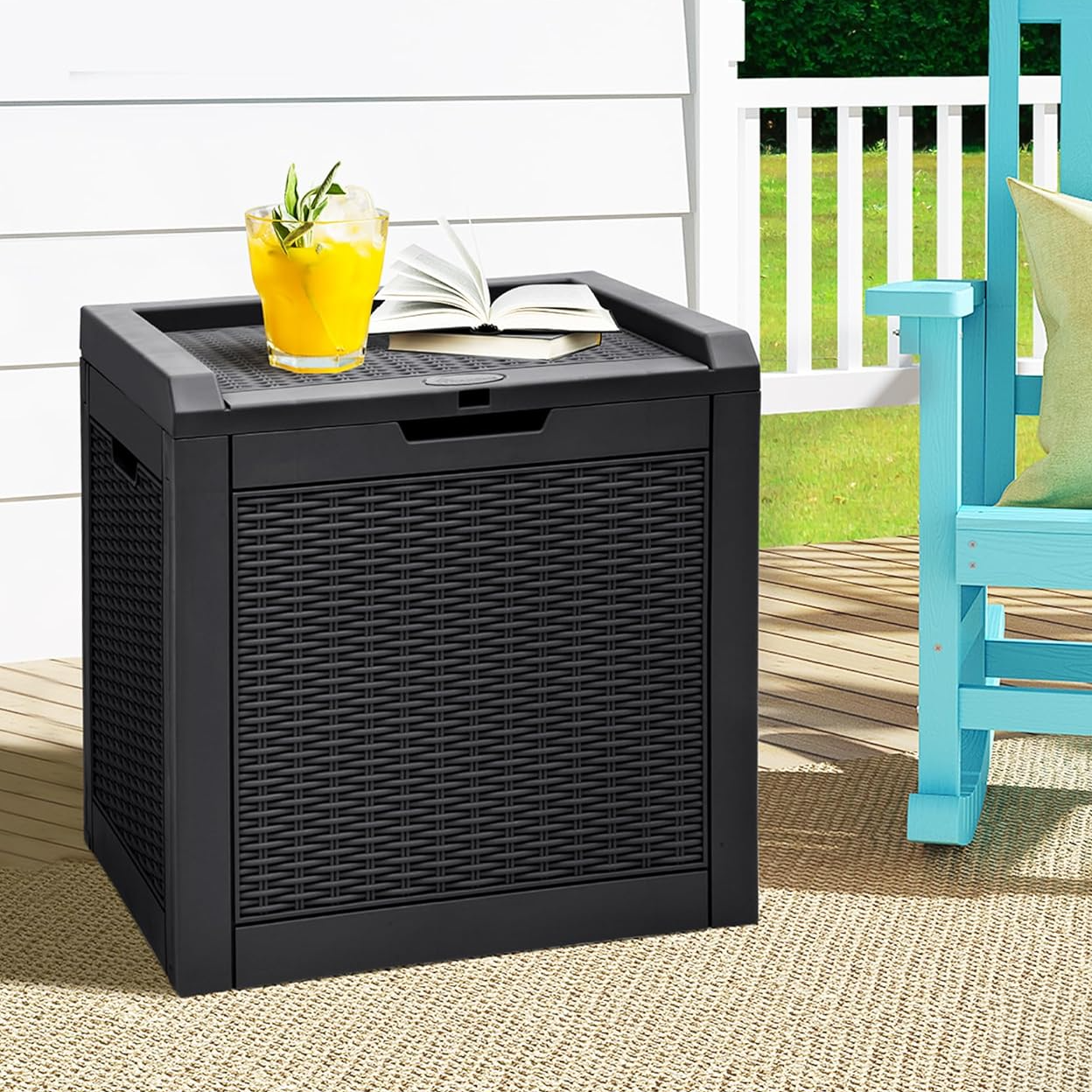 A black woven storage box serves as a tabletop with a glass of orange juice and an open book on top, situated outdoors next to a turquoise chair and a beige rug.