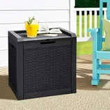 A black woven storage box serves as a tabletop with a glass of orange juice and an open book on top, situated outdoors next to a turquoise chair and a beige rug.