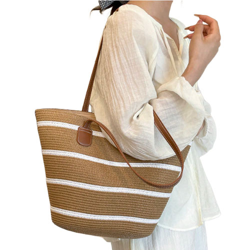 A woman is carrying a straw tote bag with brown and white stripes, featuring leather straps, while wearing a light, flowy, long-sleeve blouse.