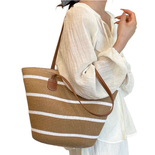 A woman is carrying a straw tote bag with brown and white stripes, featuring leather straps, while wearing a light, flowy, long-sleeve blouse.