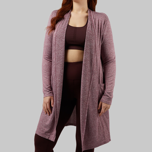 A woman is wearing a long-sleeve, knee-length, marled wine-colored wrap with an open front design. She also has on a matching sports bra and leggings.