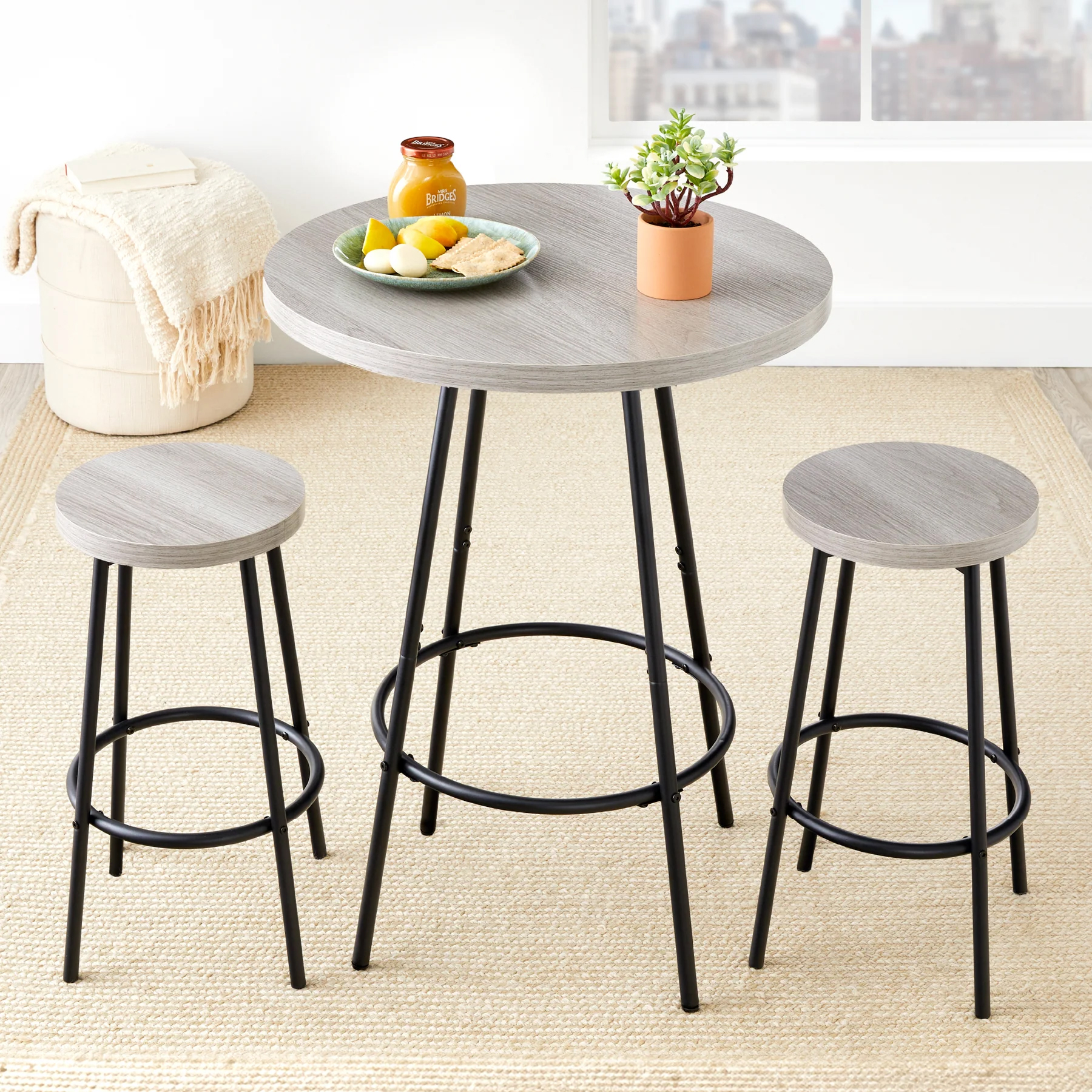 A round, gray wood-top pub table with metal legs and two matching backless stools, featuring footrests. A small plant, jar, and plate of lemons and food items are on the table, set in a room with a beige rug and a soft, textured pouf.