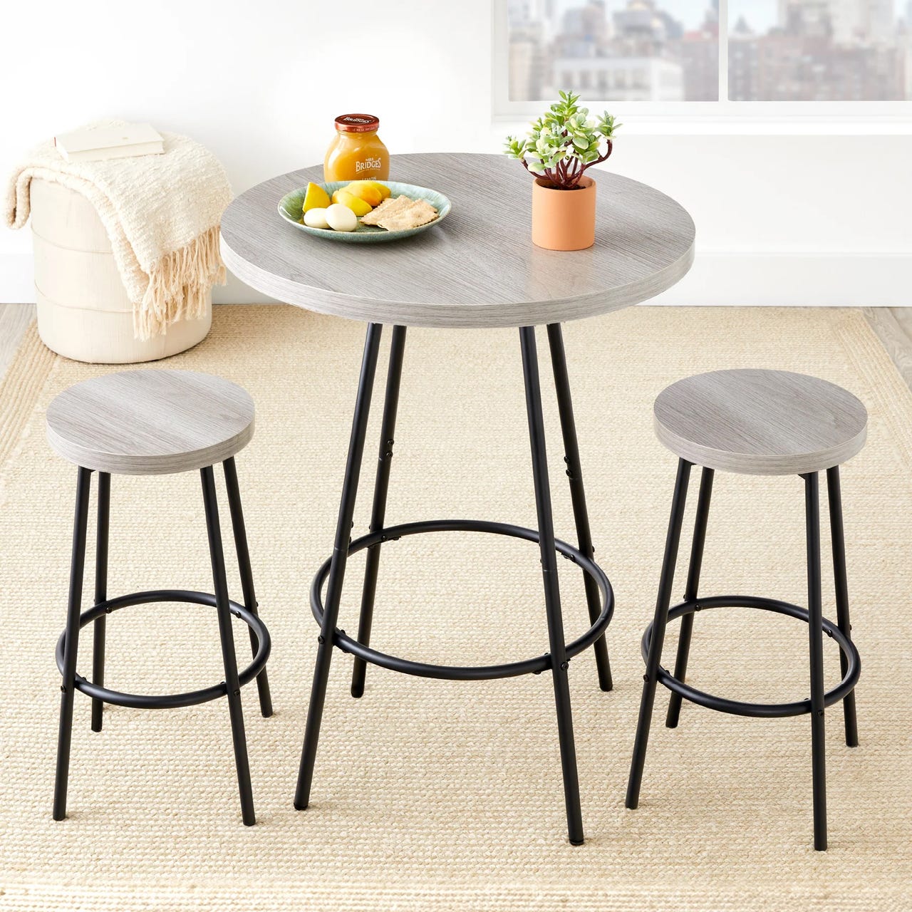 A round, gray wood-top pub table with metal legs and two matching backless stools, featuring footrests. A small plant, jar, and plate of lemons and food items are on the table, set in a room with a beige rug and a soft, textured pouf.