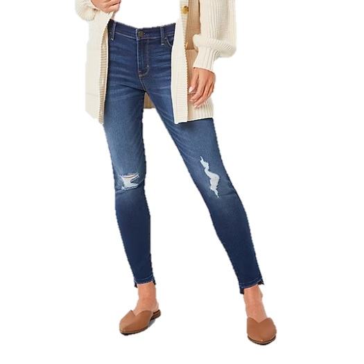 A person wearing distressed blue skinny jeans paired with a beige cardigan and brown slip-on shoes.