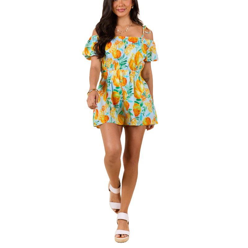 A woman is wearing an off-shoulder romper featuring a colorful citrus fruit print, paired with white platform sandals.
