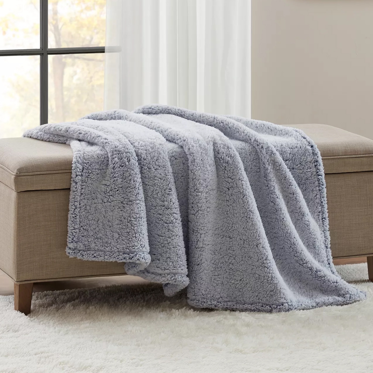 A soft, light gray fleece blanket draped over a beige upholstered bench, placed on a white plush rug near a window.