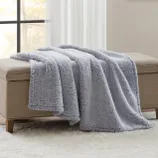 A soft, light gray fleece blanket draped over a beige upholstered bench, placed on a white plush rug near a window.