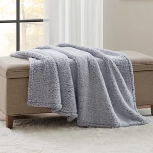 A soft, light gray fleece blanket draped over a beige upholstered bench, placed on a white plush rug near a window.