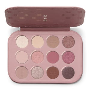 An eyeshadow palette with twelve shades labeled as Dew Drops, Moonflower, Whimsical, Belle, Vintage Peony, Shimmering Petals, Rustic Rose, Full Bloom, Dusty Lilac, Romance, Fall Petals, and Gothic Garden, housed in a pink case with the brand name UBC.