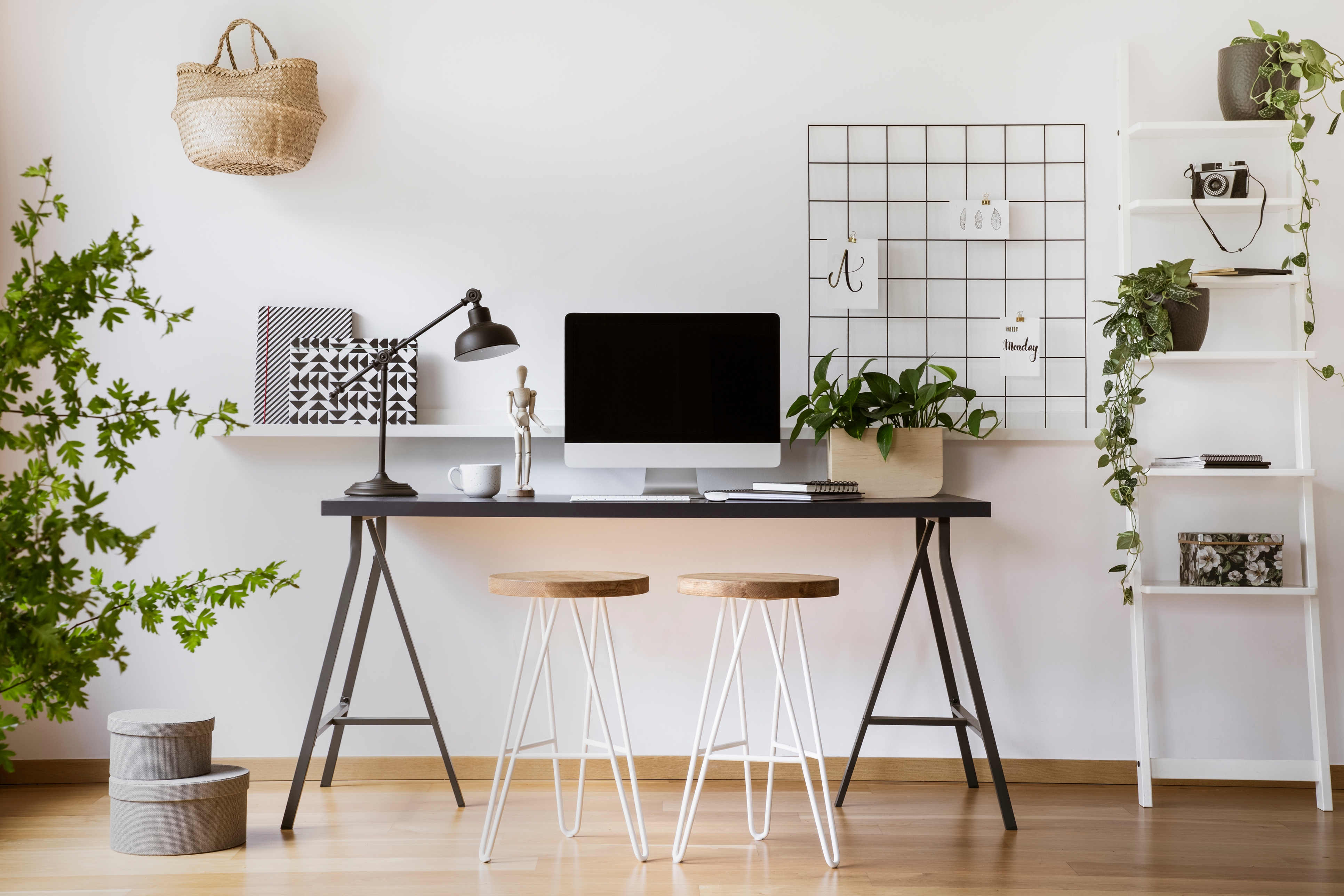 A home office setup with a computer on a desk, a desk lamp, two stools, and shelving with plants and decor items.