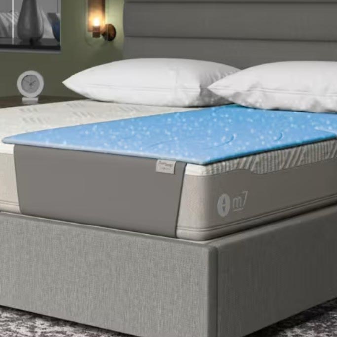A mattress topped with a blue, textured cooling layer is shown on a bed frame with a neutral upholstered base. Two pillows and part of a bedside table with a clock are visible in the background.