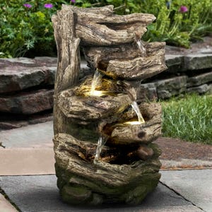 A small, tiered outdoor water fountain resembling natural stone and wood with cascading water and built-in LED lights, set on a patio with stone tiles and grass surroundings.