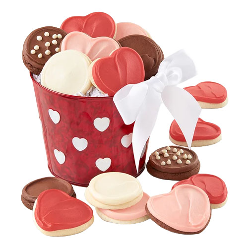 A red bucket with heart patterns is filled with assorted heart-shaped cookies in pink, white, and brown, accented with a white bow.
