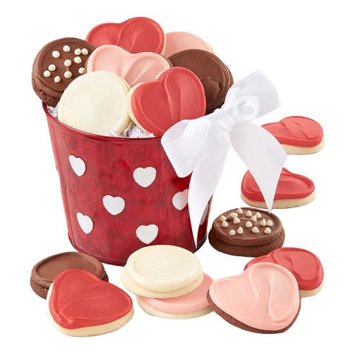 A red bucket with heart patterns is filled with assorted heart-shaped cookies in pink, white, and brown, accented with a white bow.