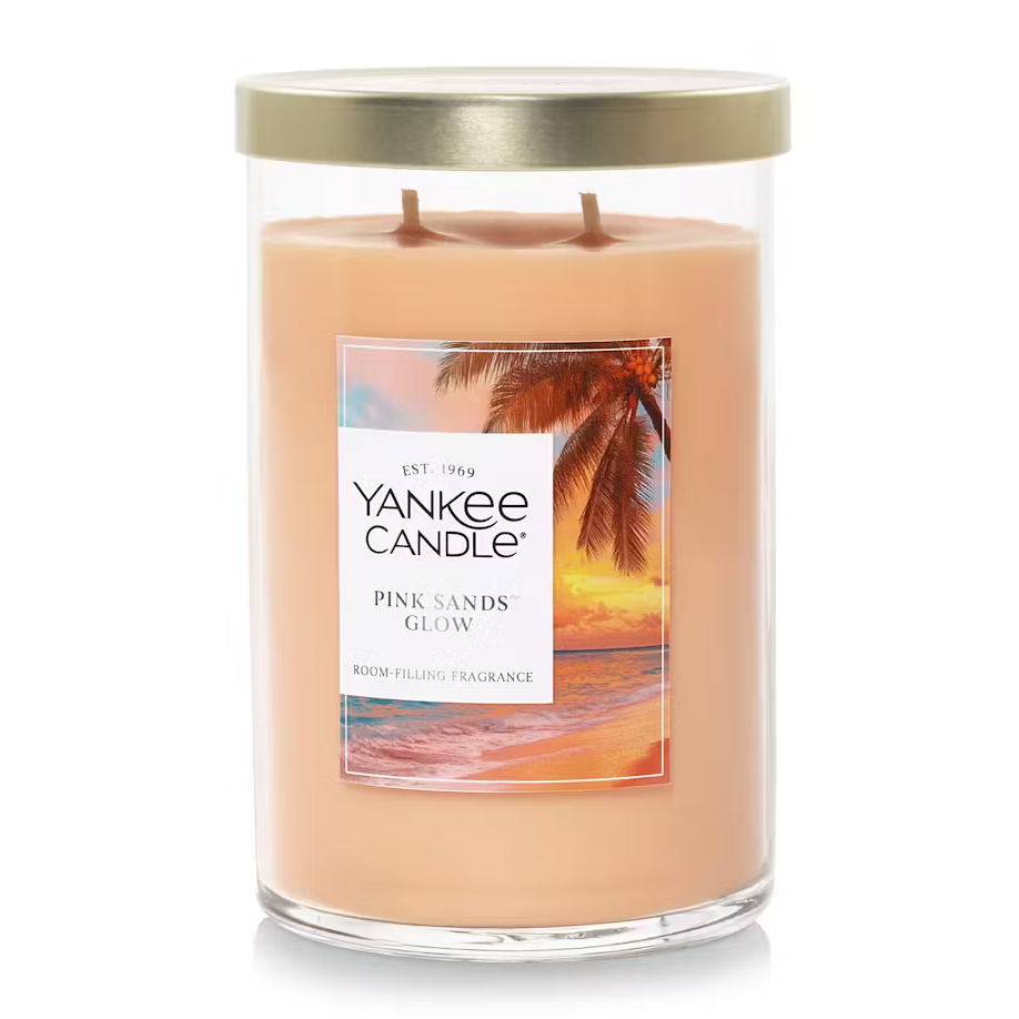 Yankee Candle's Pink Sands Glow is a two-wick candle in a clear glass jar with a metal lid, featuring a tropical beach scene label. It's designed to fill a room with its fragrance.