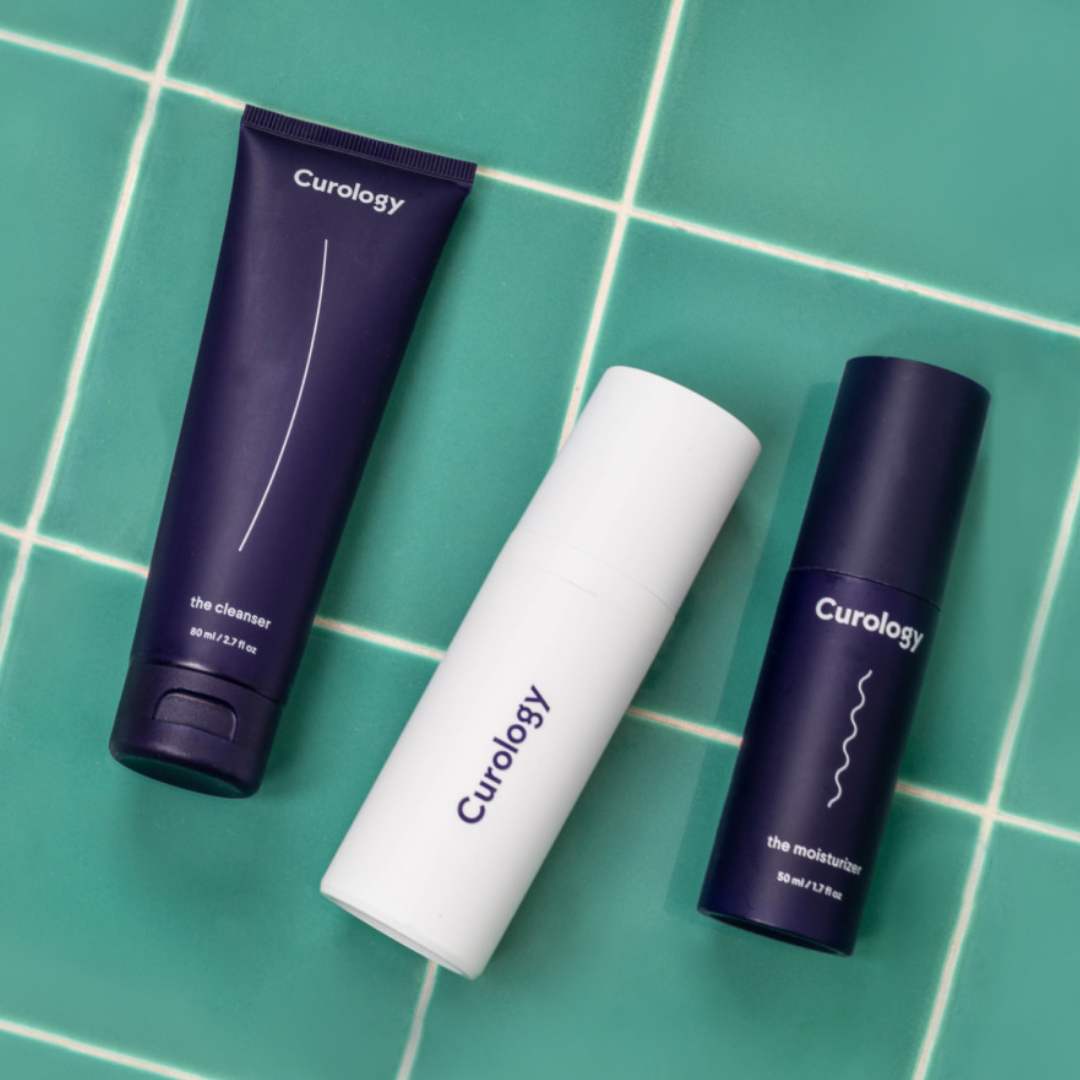 Curology skincare set on a green tiled surface: includes a cleanser (80 ml) in a dark blue tube, a white bottle, and a moisturizer (50 ml) in a dark blue bottle.