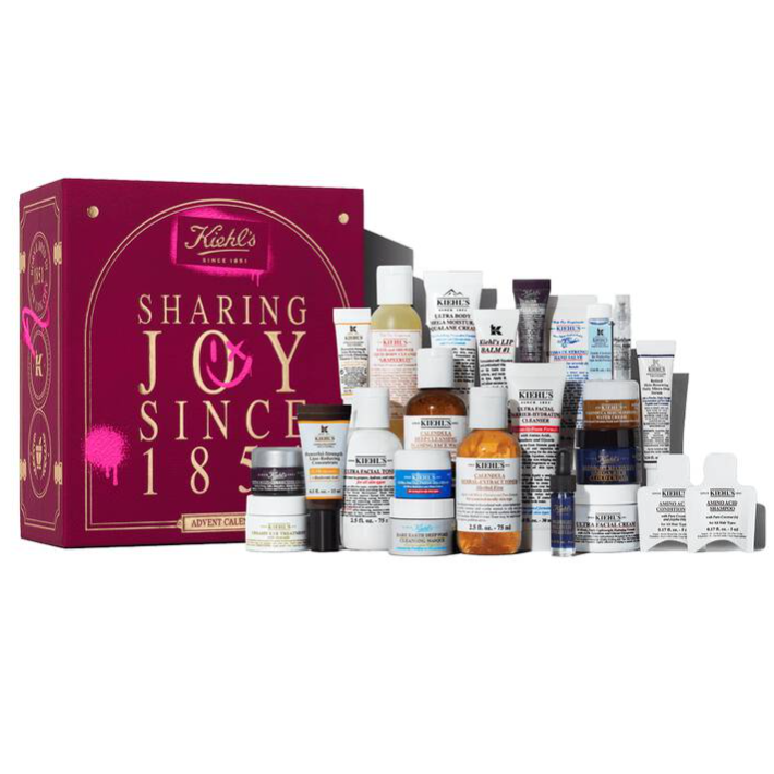 Kiehl's advent calendar featuring a variety of skincare products, including facial creams, lotions, and serums in travel sizes, displayed in front of a decorative box with \“Sharing Joy Since 1851\“ written on it.