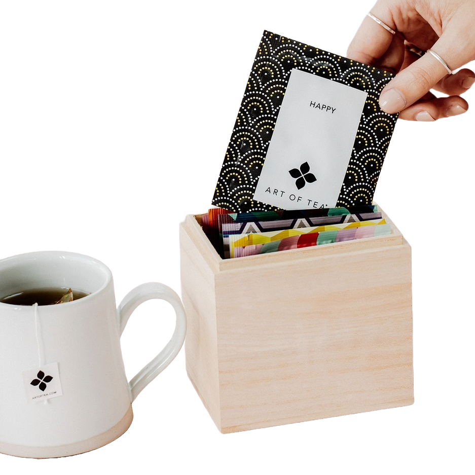 A hand is placing an \“Art of Tea\“ packet labeled \“Happy\“ into a wooden box filled with colorful tea packets. A white mug with a tea bag labeled \“Art of Tea\“ is next to the box.