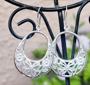 Silver dangle earrings with intricate floral and leaf cut-out design, featuring a teardrop shape, hanging from simple hooks on a decorative black stand.