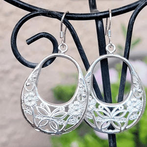 Silver dangle earrings with intricate floral and leaf cut-out design, featuring a teardrop shape, hanging from simple hooks on a decorative black stand.
