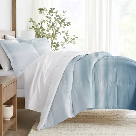 A neatly made bed features a white and light blue ombre comforter with matching pillowcases, set against a wooden headboard. A potted plant and soft natural light create a serene ambiance.