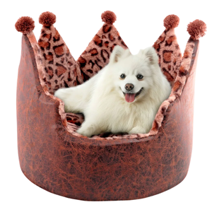 A fluffy white dog is sitting comfortably in a plush, crown-shaped pet bed with brown and leopard print details, topped with pom-poms.
