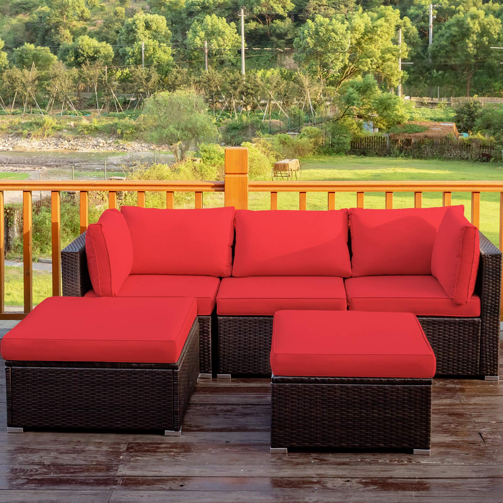 Outdoor patio set with red cushions features a modular wicker sectional sofa and two ottomans placed on a wooden deck, overlooking a lush, green landscape.