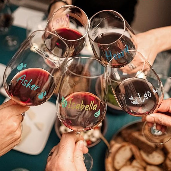 Five wine glasses with red wine being clinked together. Each glass has a name written on it: Asher, Isabella, Hui, Leo, and one partially visible name.