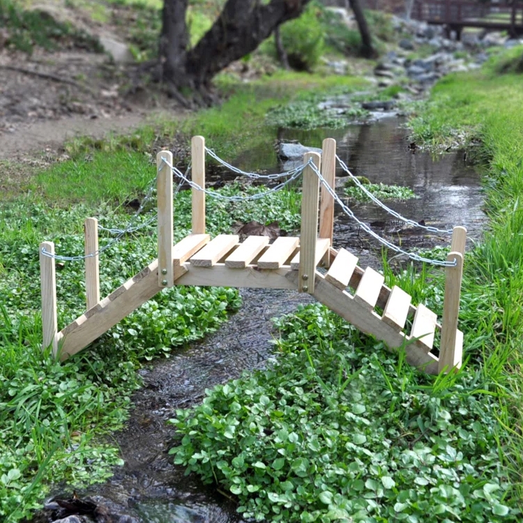 A small 3-foot wooden garden bridge with handrails and a simple slatted design spans a narrow stream, surrounded by lush green plants. It features rope chains connecting to vertical posts for added detail.